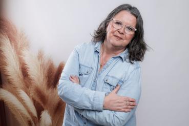 Das Bild zeigt Jutta. Sie trägt eine hellblaue Jeansjacke, eine goldene Halskette und eine weiße Brille. Ihre Arme sind vor der Brust verschränkt, und sie blickt direkt in die Kamera. Im Hintergrund ist verschwommenen Pampasgras zu sehen.