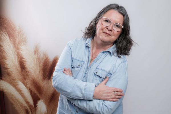 Das Bild zeigt Jutta. Sie trägt eine hellblaue Jeansjacke, eine goldene Halskette und eine weiße Brille. Ihre Arme sind vor der Brust verschränkt, und sie blickt direkt in die Kamera. Im Hintergrund ist verschwommenen Pampasgras zu sehen.